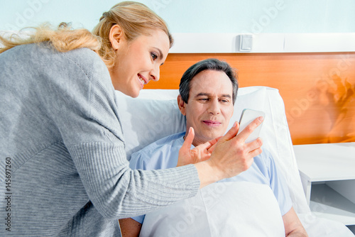 wife visiting her middle aged husband and showing him smartphone in hospital