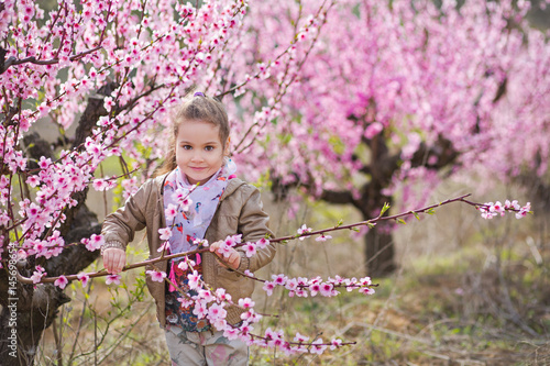 Cute beautiful stylish dressed blond girl standing on a field of spring young peach tree with pink flowers.smiling girl dressed in in pink in spring season.