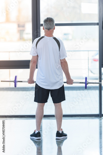 Back view of mature man in sportswear holding barbell in gym