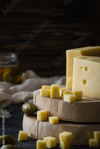 Swedish hard yellow cheese with holes chopped on wooden slices with green olives on dark rustic background