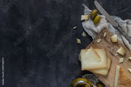 Italian hard cheese pecorino toscano sliced and chopped on wooden board with knife and green olives on dark rustic background, top view with copyspace
