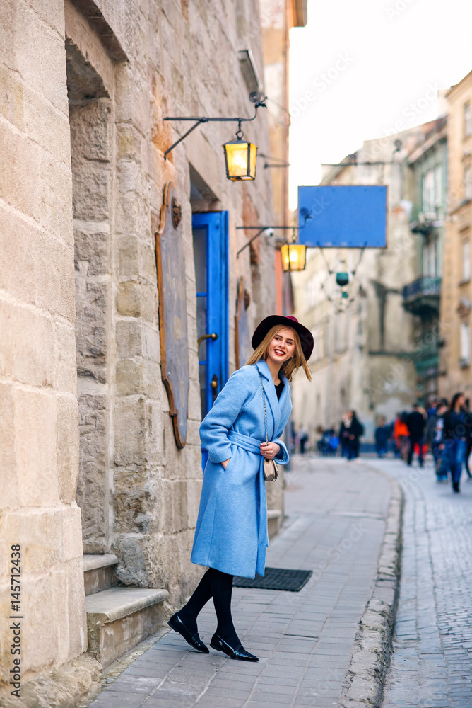 Beautiful young tourist girl in a blue coat and burgundy hat standing in front of the cozy cafe with blue doors and vintage lanterns. Attractive girl with perfect smile.