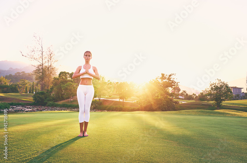 Yoga at park with view of the mountains, with sunlight. Young woman in asana pose standing on green grass. Concept of calm and meditation.