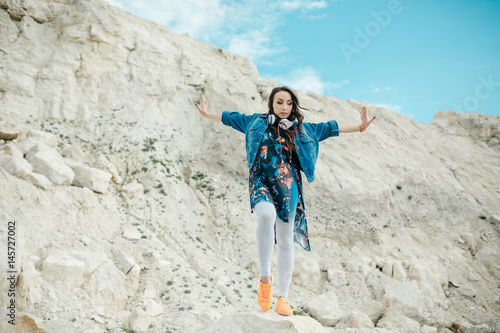 Young woman with headphones posing and dancing near the beautiful mountains wearing colorful clothes