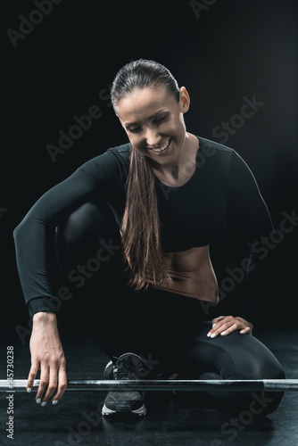 young fitness woman sitting at barbell and resting isolated on black.