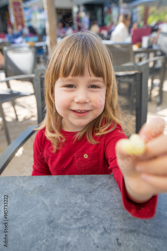 portrait of blonde three years old child face, with red shirt, sitting in terrace exterior bar cafe with grey table, offering cheese puff in hand 