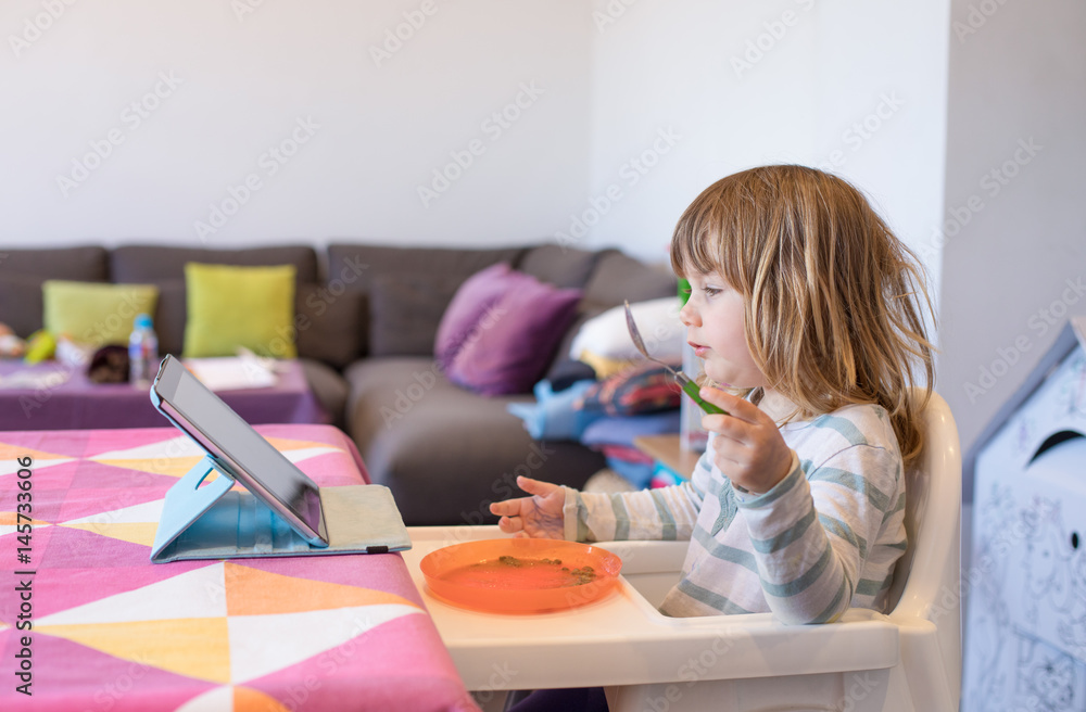 Three years old blonde child sitting in white high chair indoor, eating from orange plastic dish, and watching digital tablet on the table
