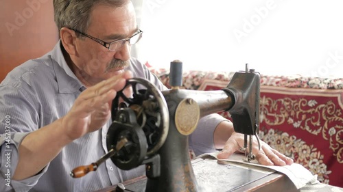 An adult man with a mustache sews on an old hand-sewn machine. Glasses are dressed and white cloth is sewed on.