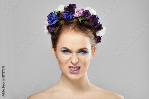 Young angry girl with flowers in her hair grins. Perfect creative make up and floral art hairstyle. photo