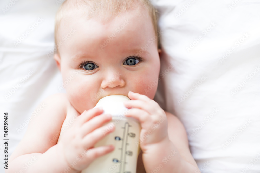 Pretty baby girl drinks water from bottle lying on bed. Child weared diaper in nursery room. Pretty baby girl drinks water from bottle lying on bed. Child weared diaper in nursery room.