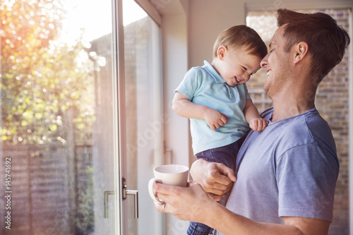 Father holds toddler son while drinking coffee at home, by the window