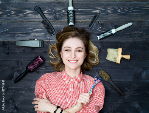 Happy smiling young woman in plaid shirt with scissors in hand and hairdresser tools among her. Top view