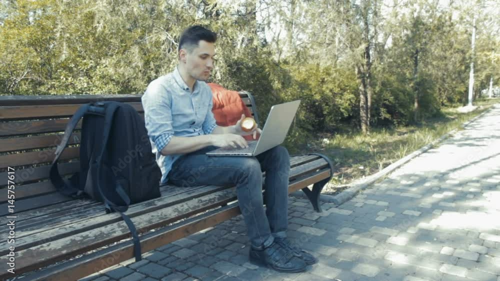 Young man in shirt siting on the bench in green park and typing on his laptop and eating apple Young man in shirt siting on the bench in green park and typing on his laptop and eating apple