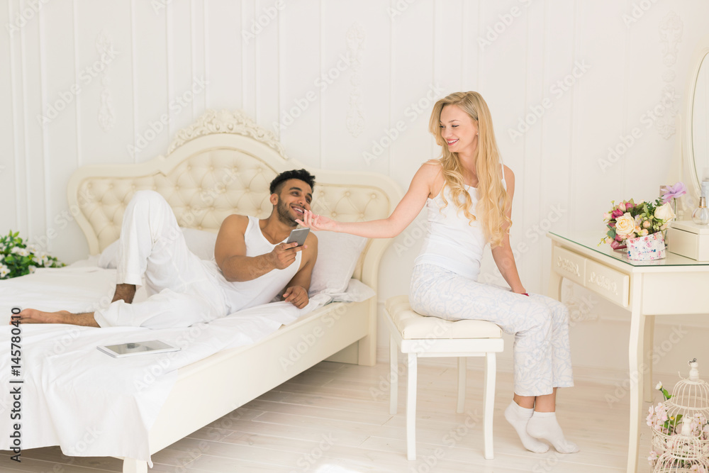 Young Couple In Bedroom, Hispanic Man Using Cell Smart Phone Lying Bed And Woman Sitting Happy Smile Loves Young Couple In Bedroom, Hispanic Man Using Cell Smart Phone Lying Bed And Woman Sitting Happy Smile Loves