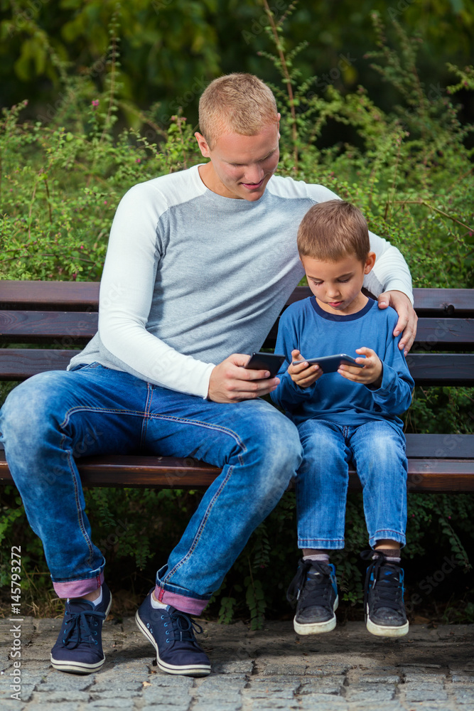 Fototapeta premium Young father and his little son are using mobile phones while relaxing on a park bench