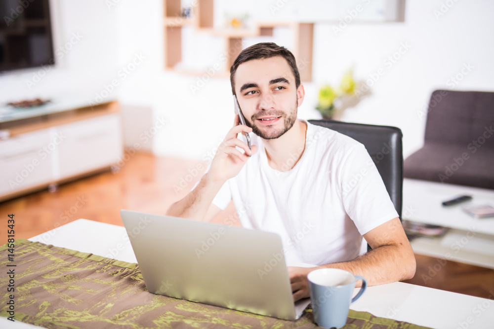 Handsome businessman is talking on the mobile phone and smiling while using a laptop in kitchen Handsome businessman is talking on the mobile phone and smiling while using a laptop in kitchen