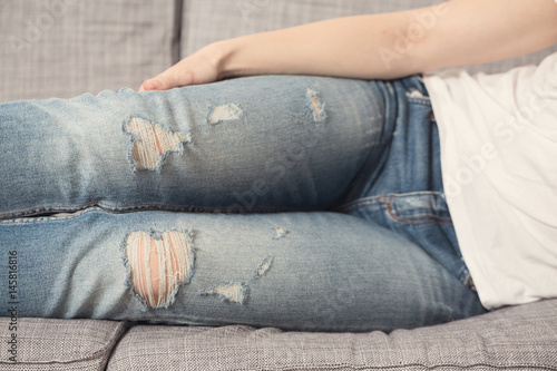 Girl with denim fabric pants, sitting on the sofa; denim torn on the knees and threads coming apart
