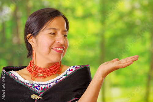 Closeup beautiful hispanic woman wearing traditional andean white blouse with colorful decoration around neck, black poncho, matching red necklace and ear, posing happily for camera, garden background