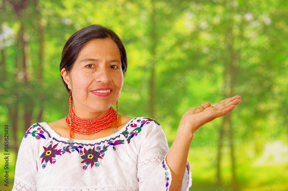 Closeup beautiful hispanic woman wearing traditional andean white blouse with colorful decoration around neck, matching red necklace and ear, posing happily for camera holding out palm of hand, garden Closeup beautiful hispanic woman wearing traditional andean white blouse with colorful decoration around neck, matching red necklace and ear, posing happily for camera holding out palm of hand, garden