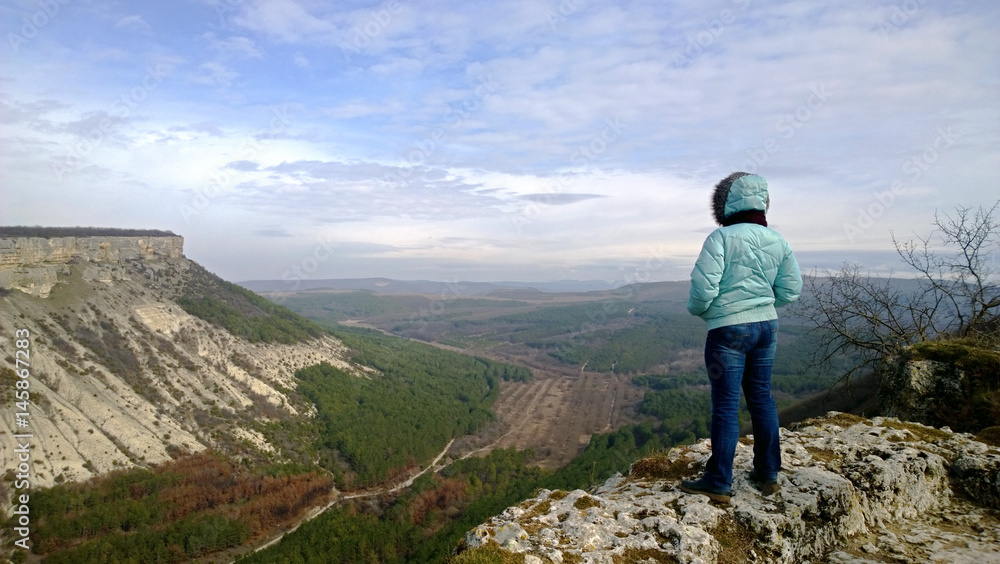Lonely Mature woman standing on edge of cliff and looks at view. Dressed in a jacket and jeans. The view from the back. Windy and cool