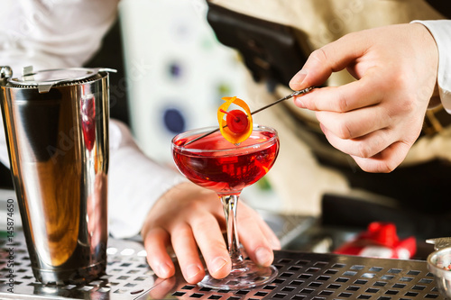 Bartender is holding orange rind in tongs decorating red cocktail at bar background.