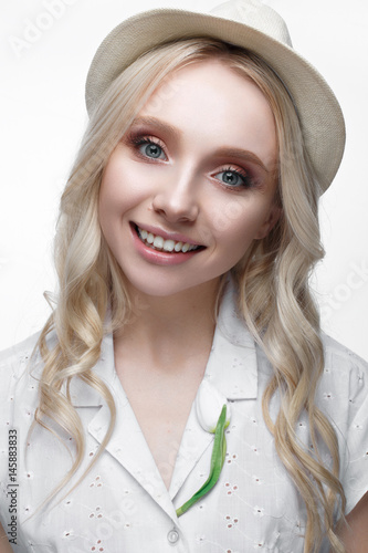 Young smiling girl with curls in a hat. A beautiful model in a white shirt and a brooch-flower. Gentle makeup. Summer light image. Beauty of the face. photo