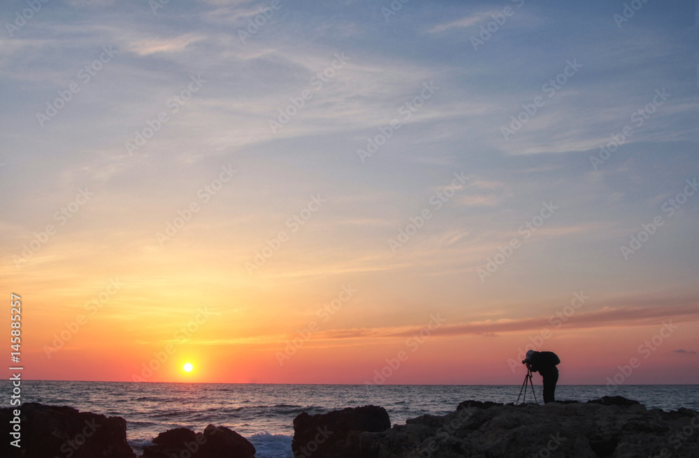 Silhouette of a photographer with a backpack and tripod with a beautiful sea sunset on the background