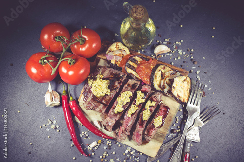 Rare roast steak of beef with sharp green mustard. Grilled vegetables and spices on rustic chopping board on dark background. Food background. Selective focus. Top view
