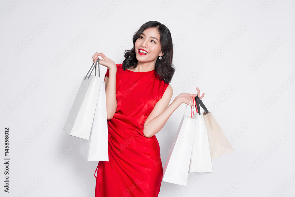 Cheerful fashionable asian woman wearing a red dress with shopping bags standing over white. Cheerful fashionable asian woman wearing a red dress with shopping bags standing over white.