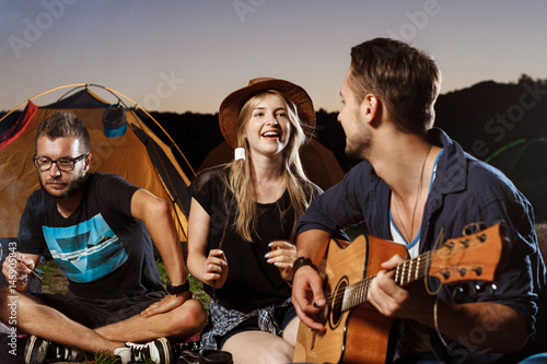 Friends sitting near bonfire, smiling, playing guitar. Camping grill marshmallow. © Cookie Studio