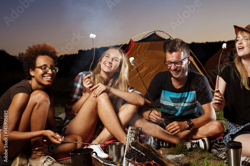 Friends sitting near bonfire, smiling, playing guitar. Camping grill marshmallow. © Cookie Studio