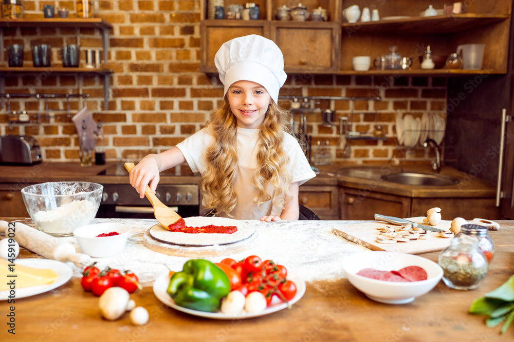 smiling girl in chef hat putting tomato sauce on pizza dough