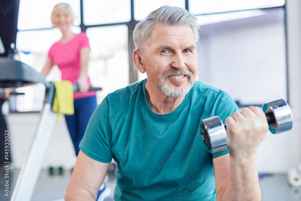 senior sportsman sitting and training with dumbbell, sportswoman on treadmill behind. senior fitness class concept senior sportsman sitting and training with dumbbell, sportswoman on treadmill behind. senior fitness class concept