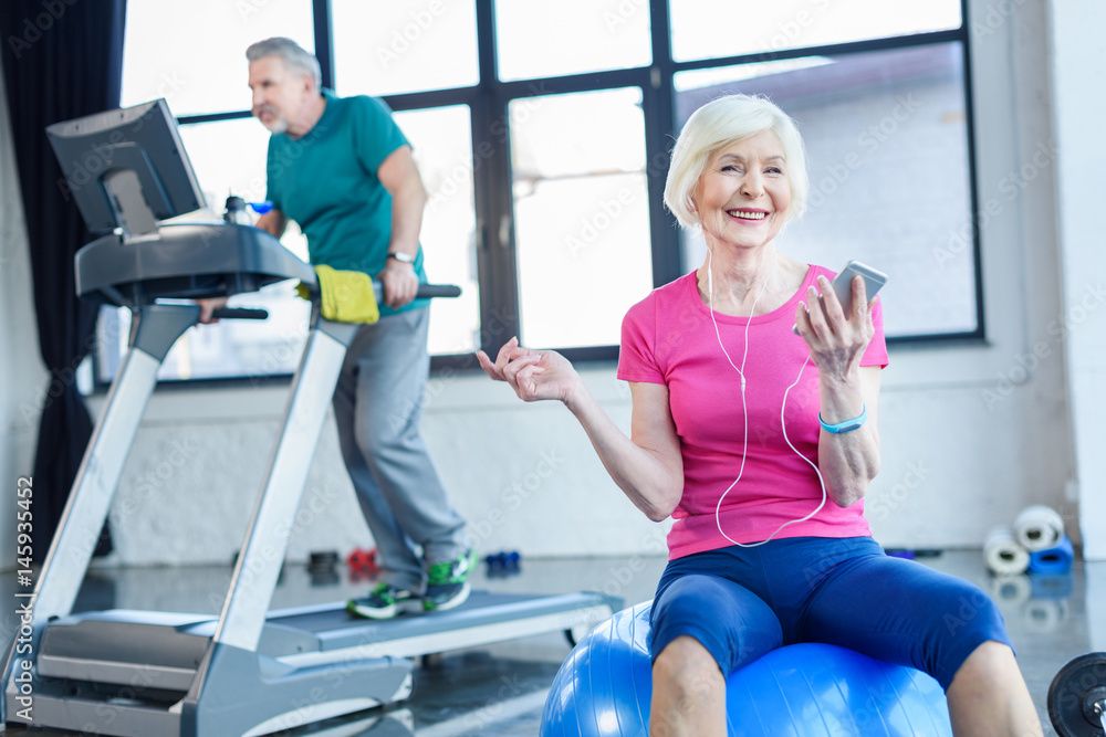 senior sportswoman sitting on fitness ball with smartphone, sportsman on treadmill behind in senior fitness class senior sportswoman sitting on fitness ball with smartphone, sportsman on treadmill behind in senior fitness class