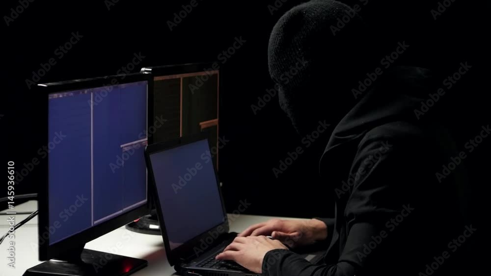 Hacker in balaclava cracking code using laptop and computers from his dark hacker room Hacker in balaclava cracking code using laptop and computers from his dark hacker room