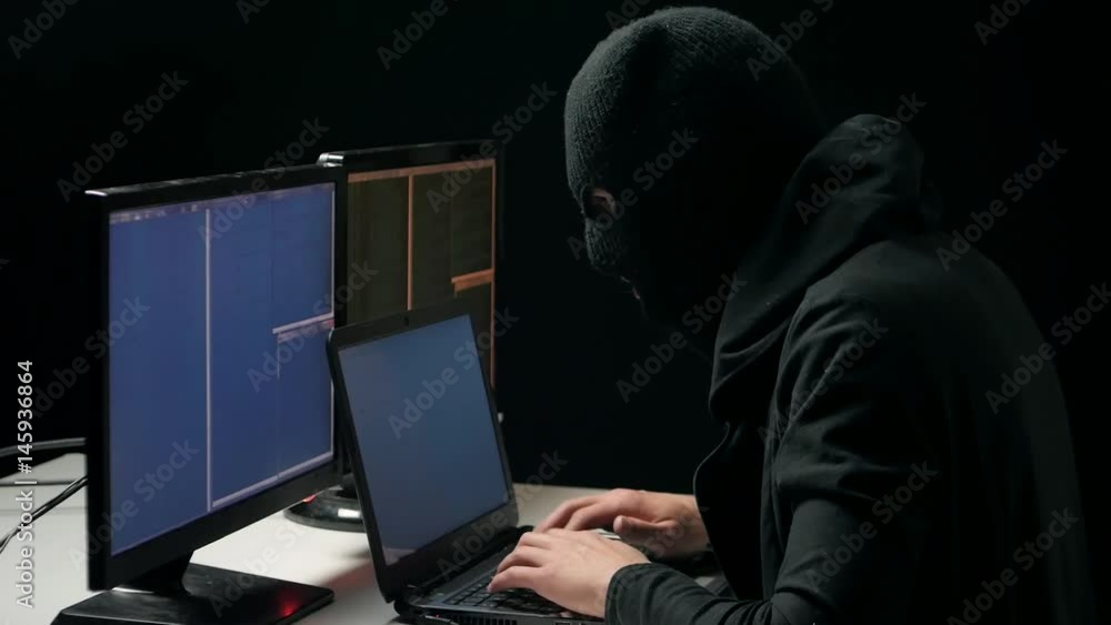 Hacker in balaclava cracking code using laptop and computers from his dark hacker room Hacker in balaclava cracking code using laptop and computers from his dark hacker room