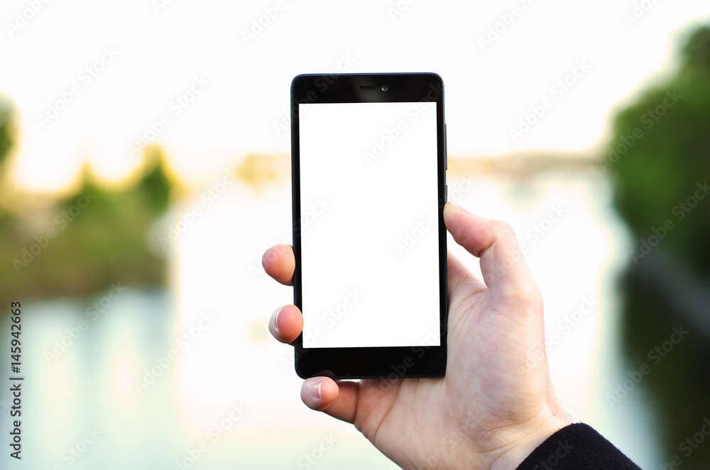 Photo of a man's hand holding black phone with blank screen with blurred lake or river in the background Photo of a man's hand holding black phone with blank screen with blurred lake or river in the background