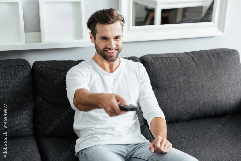 Fototapeta premium Young cheerful man sitting on sofa holding remote control.