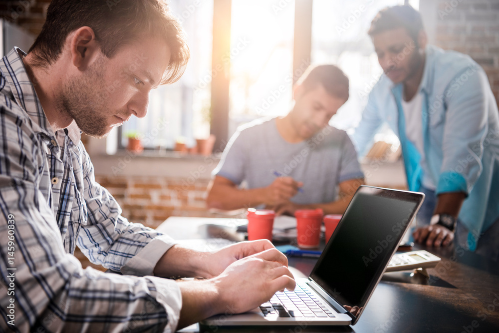 Young man using laptop while colleagues discussing project, small business meeting concept Young man using laptop while colleagues discussing project, small business meeting concept