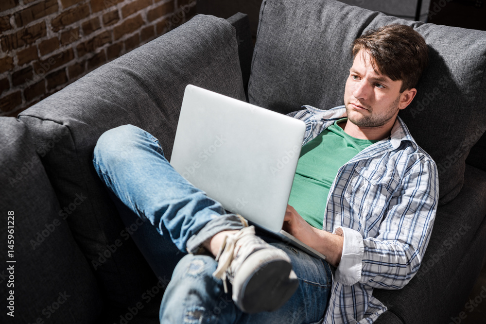 Serious young man lying on sofa and using laptop at home, small business people concept