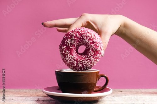 Woman hand holding colorful donut with sprinkles on a wooden table and pink background. Cup of coffee. Concept of food. Donut with bite missing Close up. Pastel color 
