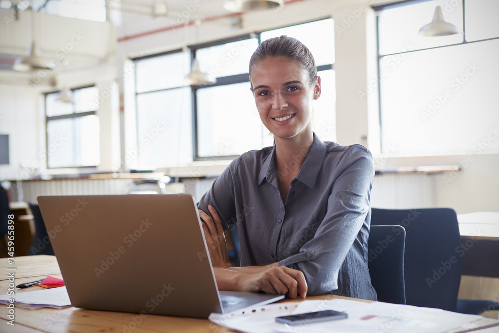 Young woman in office using laptop, smiling to camera Young woman in office using laptop, smiling to camera