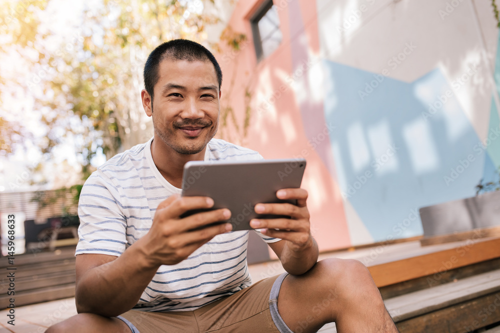 Smiling Asian man sitting on stairs outside using a tablet Smiling Asian man sitting on stairs outside using a tablet