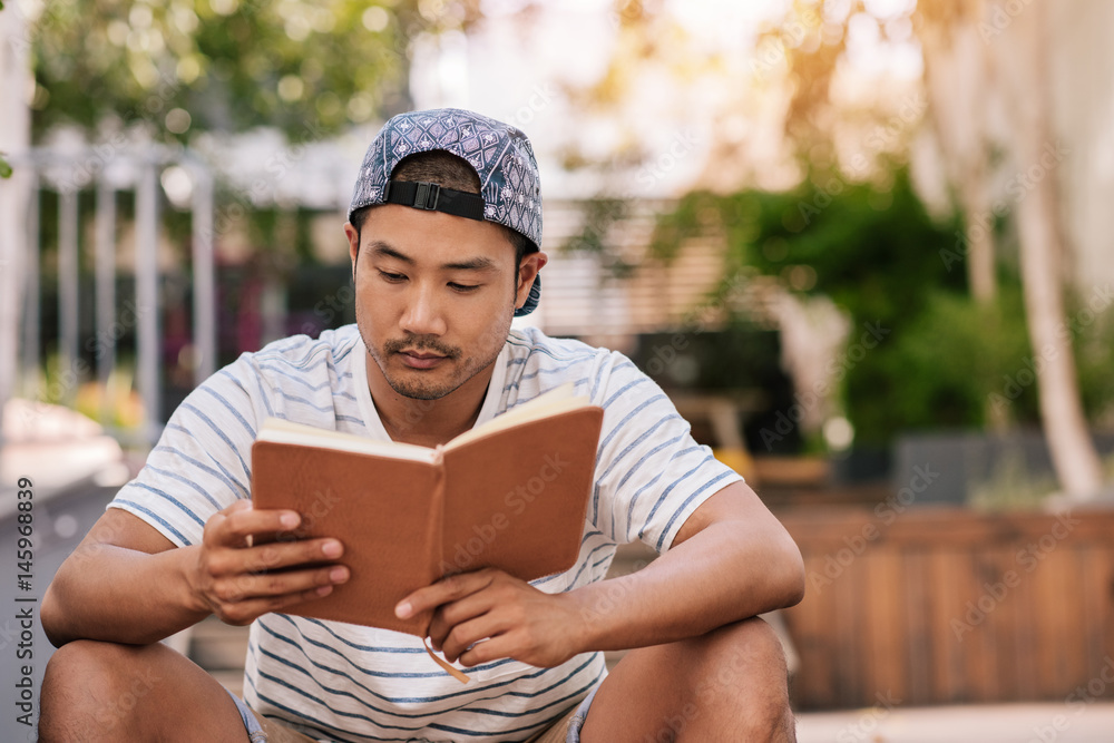 Fototapeta premium Young Asian man sitting on stairs outside reading a book