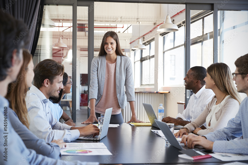 Woman addressing team leans on desk at business meeting Woman addressing team leans on desk at business meeting
