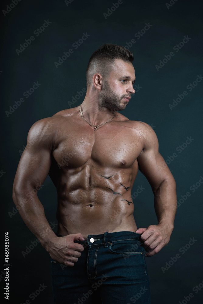 Handsome muscular well-shaped tanned sexy bodybuilder/male fitness model with tattoo and perfect abs poses over dark background looking to his left in natural colors