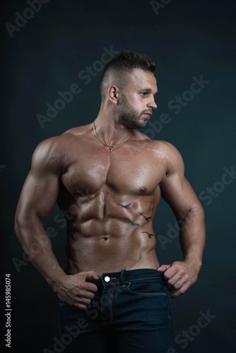 Handsome muscular well-shaped tanned sexy bodybuilder/male fitness model with tattoo and perfect abs poses over dark background looking to his left in natural colors