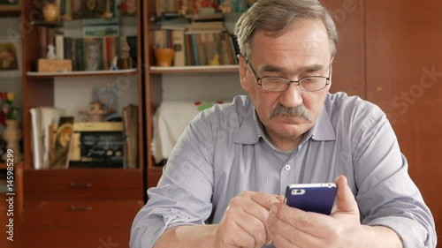 An elderly man with a mustache in glasses dials a text message on his mobile phone. Sits at home at the table.