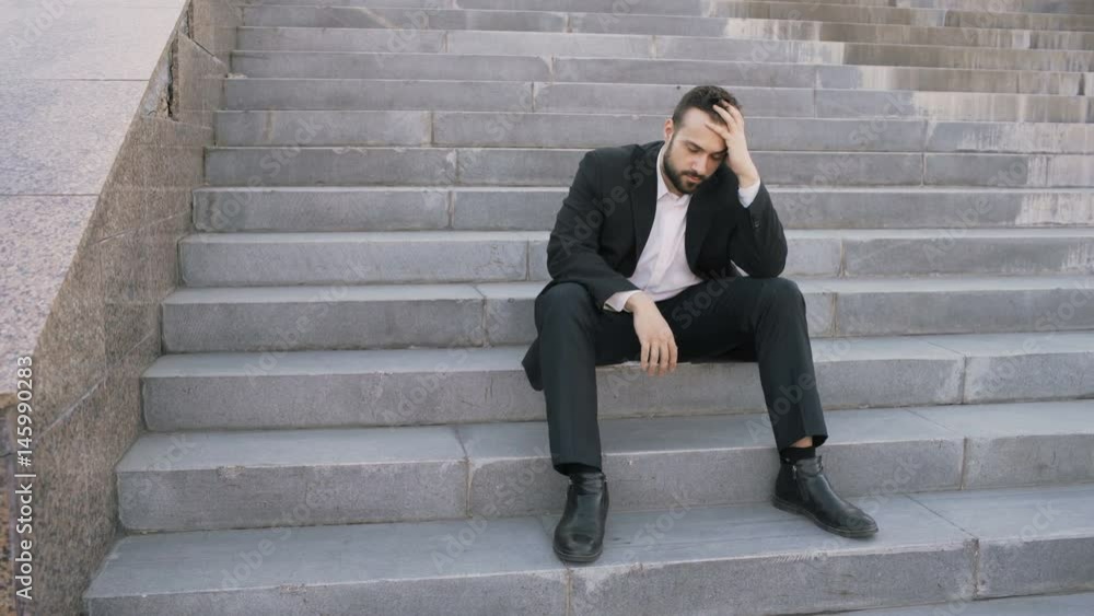 Upset young business man having stress and sitting on stairs in street. Businessman having deal problems concept Upset young business man having stress and sitting on stairs in street. Businessman having deal problems concept