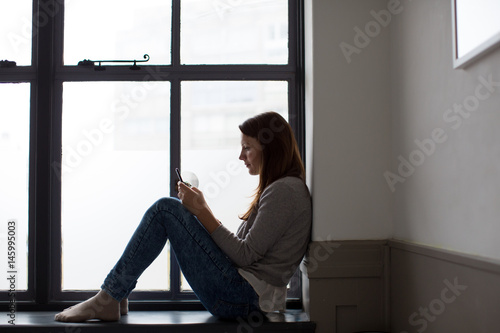 Young adult female sitting on window ledge with smartphone photo